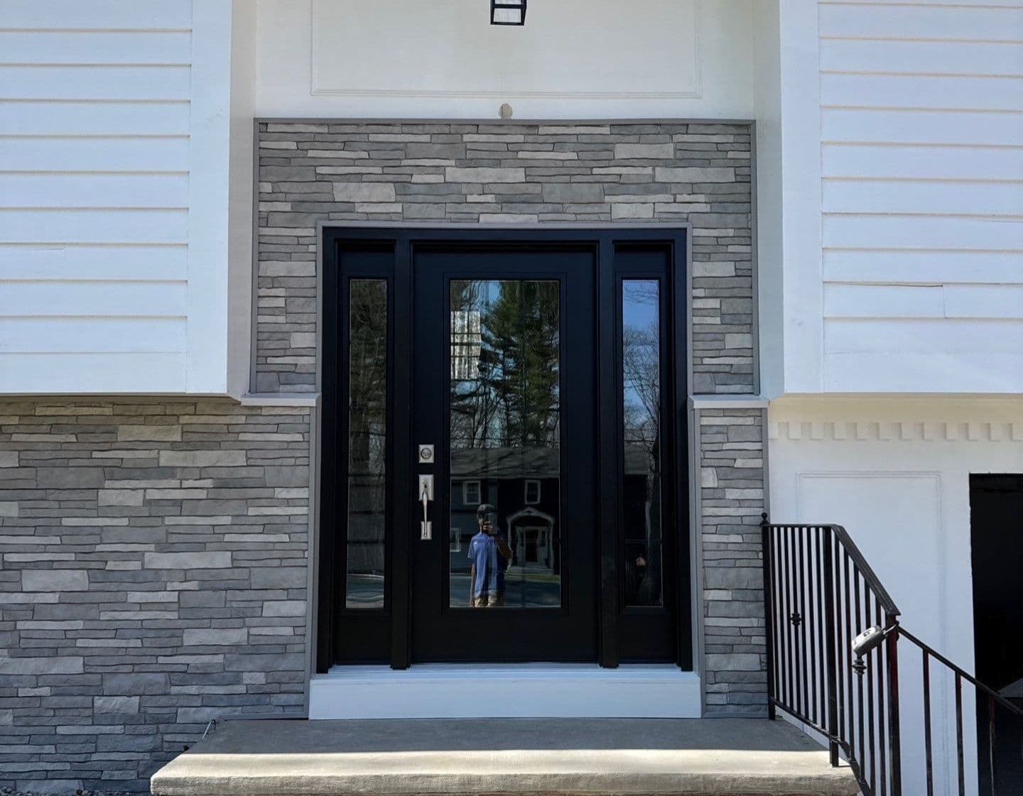 Modern home entrance with black French doors and sidelights, surrounded by gray stone veneer and white siding, pendant light fixture above, metal stair railing to right
