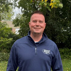 Man wearing navy quarter-zip jacket with company logo, smiling at camera with trees and garden foliage in background
